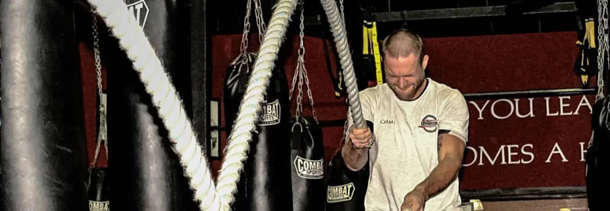 A man engaged in an intense battle rope workout at Creighton MMA, surrounded by heavy bags and training equipment in the gym.