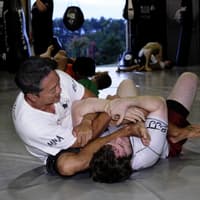 Two Brazilian Jiu-Jitsu practitioners at Creighton MMA engaged in training, with one applying an armbar submission while others drill in the background.