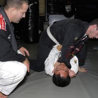 Head instructor Paul Creighton observes two students practicing a submission technique, with one attempting a choke from the top position.