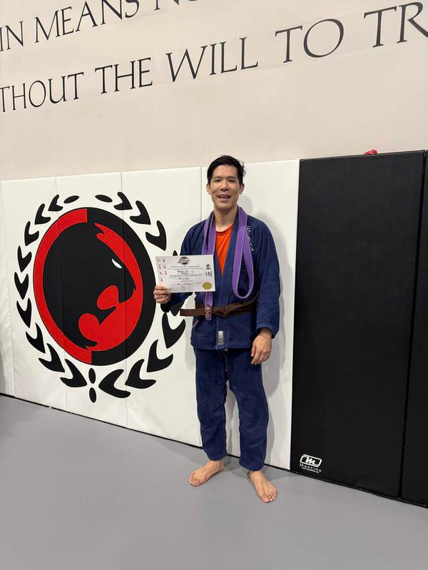 Michael Yoon in a jiu-jitsu gi holding a certificate, standing in front of a martial arts gym wall with a logo and motivational quote.